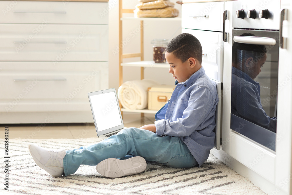 Little African-American boy with laptop studying computer sciences online in kitchen