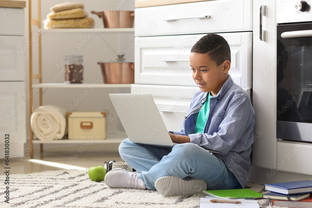 Little African-American boy with laptop studying computer sciences online in kitchen