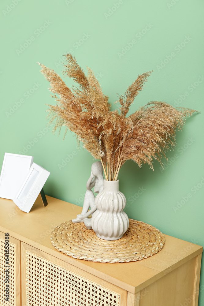 Vase of dried pampas grass with figurine and photo frames on dresser near green wall