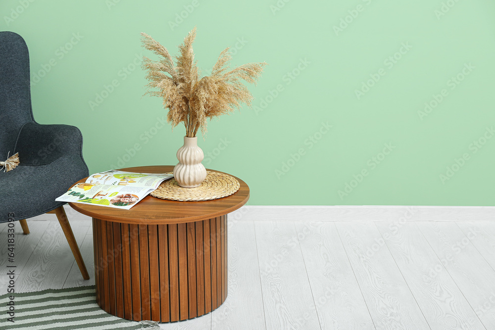 Vase of dried pampas grass with magazine on coffee table and armchair near green wall