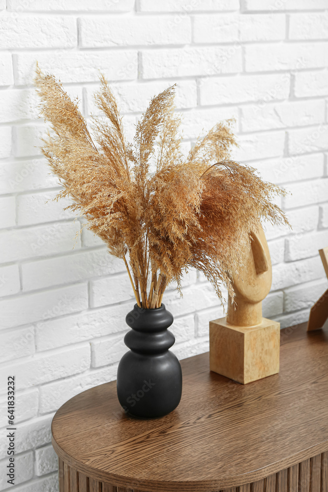 Vase of dried pampas grass with figurine on dresser near white brick wall