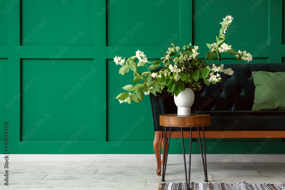 Vase with blooming jasmine flowers on coffee table and sofa near green wall