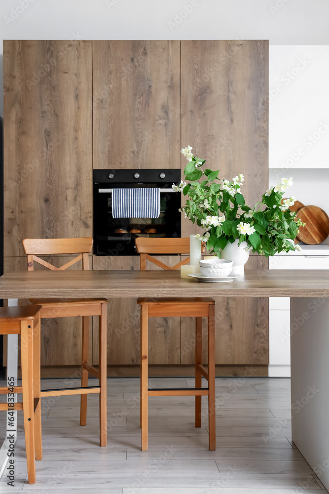 Vase with blooming jasmine flowers, candles and bowls on wooden table in modern kitchen