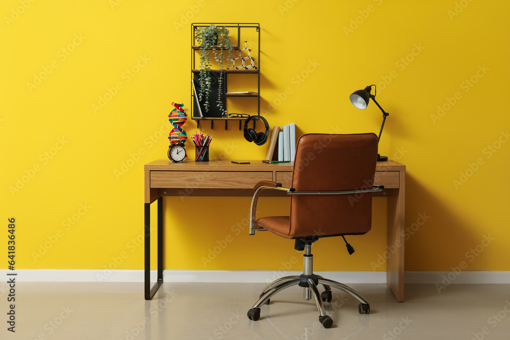 Modern school desk with molecular model, alarm clock and stationery in room near yellow wall