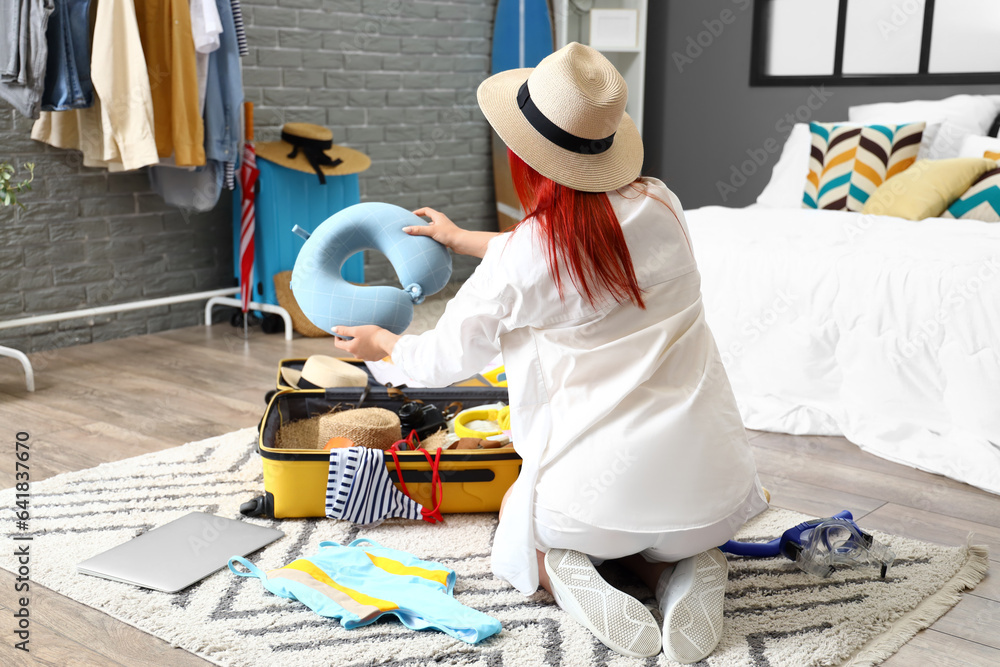 Young woman with neck pillow unpacking suitcase in bedroom