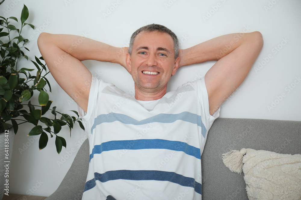 Happy mature man with hands behind his head sitting on grey sofa in living room