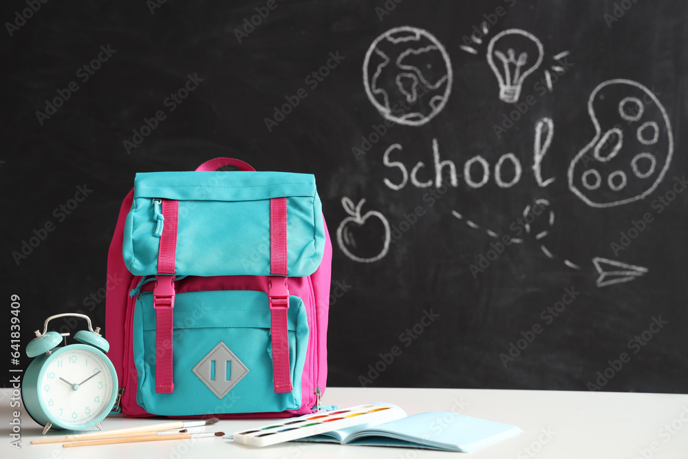 Colorful school backpack with stationery and alarm clock on white table near black chalkboard