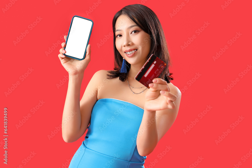 Beautiful stylish young Asian woman with mobile phone and credit card on red background