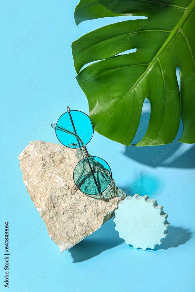 Stone with stylish sunglasses and monstera leaf on blue background