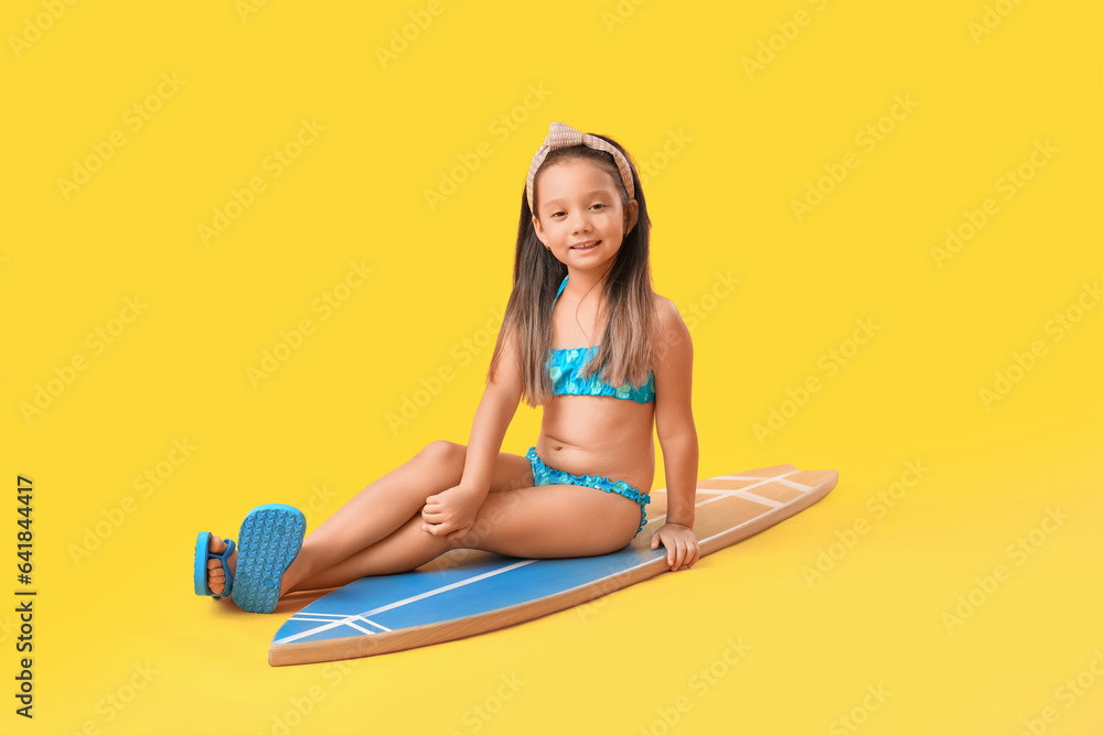 Little Asian girl with surfboard on yellow background