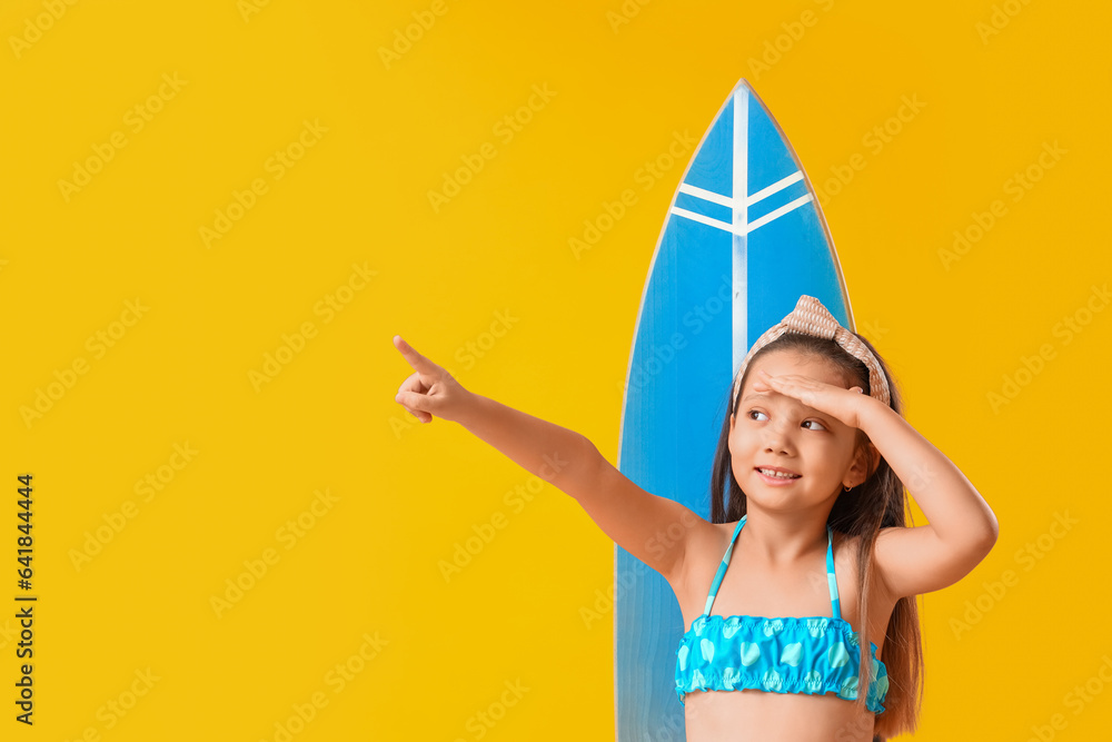 Little Asian girl with surfboard pointing at something on yellow background