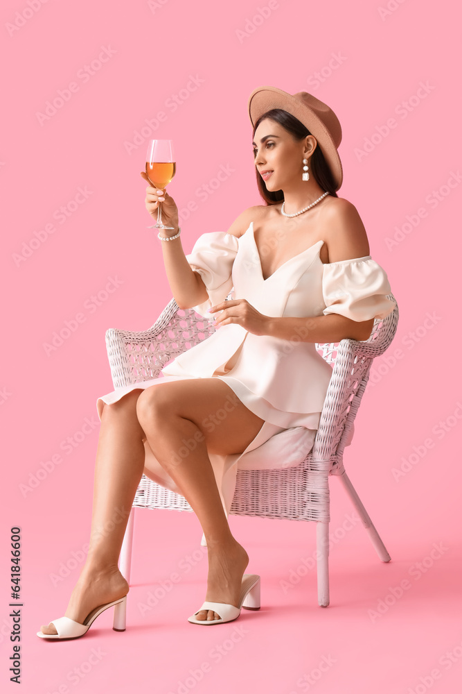 Young woman with glass of wine sitting in armchair on pink background