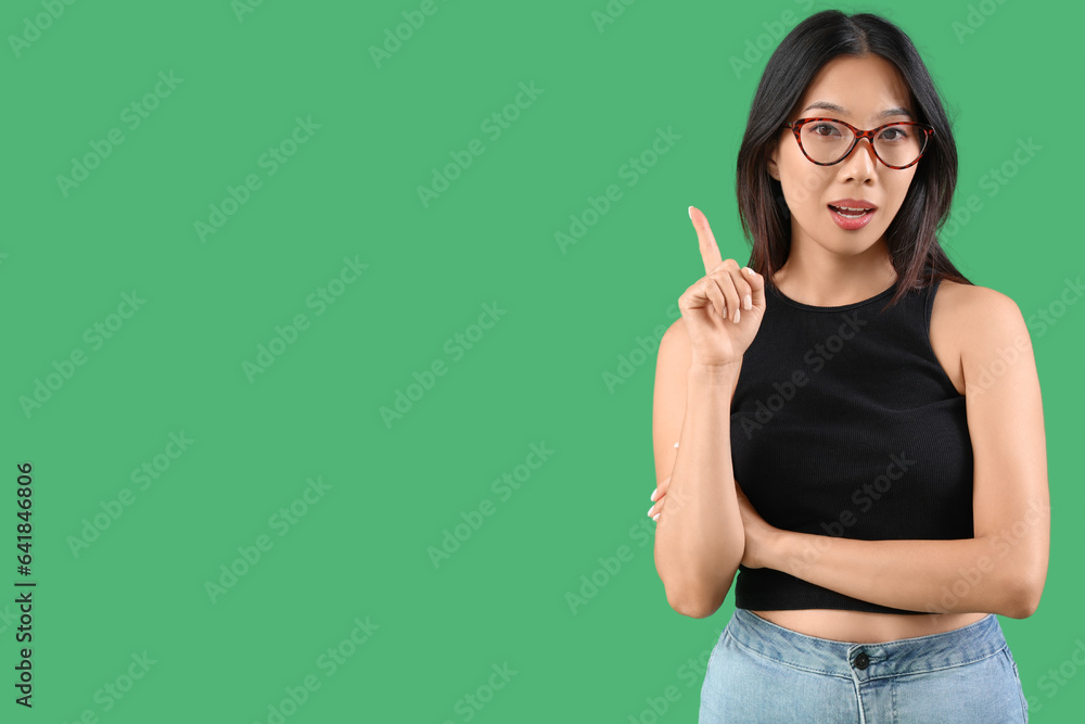 Thoughtful young beautiful Asian woman in stylish eyeglasses pointing at something on green backgrou