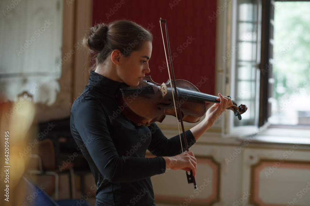 In a classical setting, a lovely young woman performs on the violin by the window