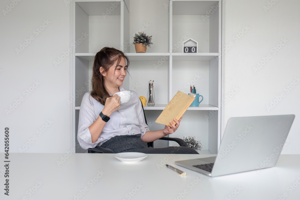 Young office worker sitting at table drinking coffee while checking documents and working on laptop.