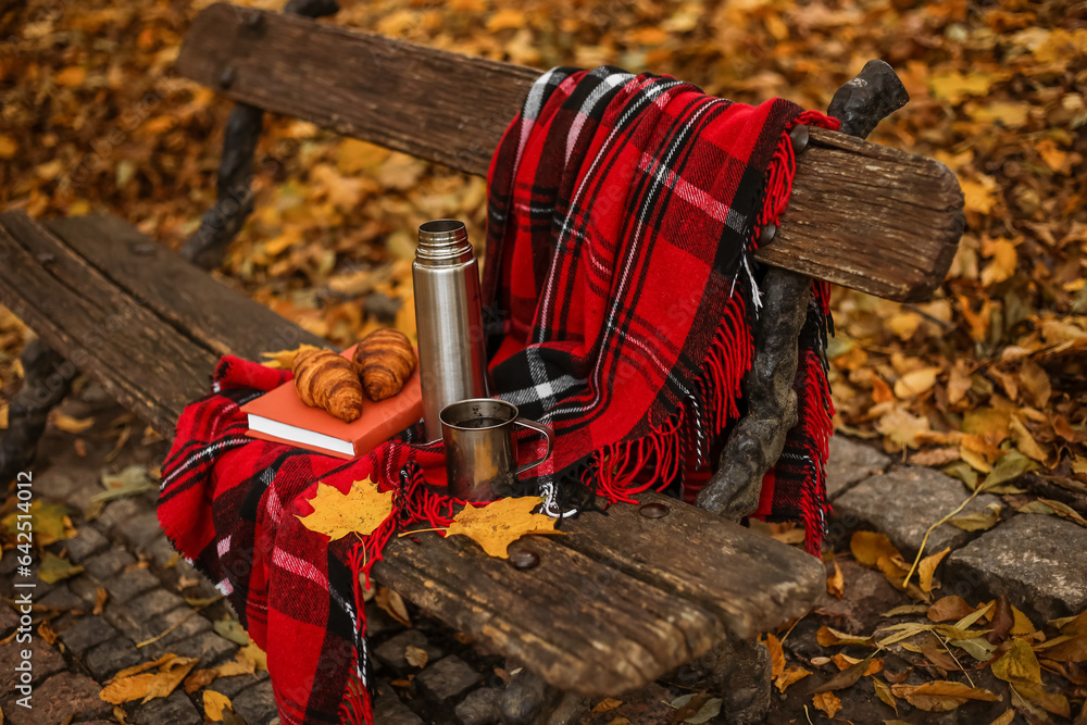 Book with croissants, thermos, cup of tea and plaid on bench in autumn park