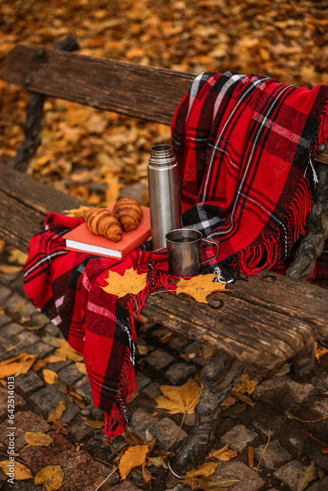 Book with croissants, thermos, cup of tea and plaid on bench in autumn park