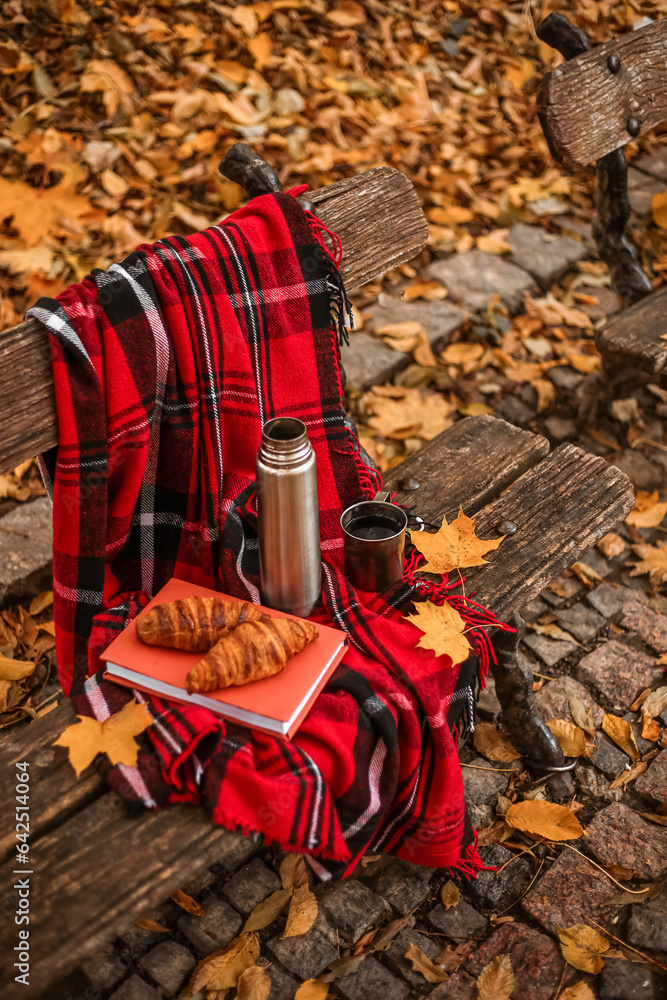 Book with croissants, thermos, cup of tea and plaid on bench in autumn park