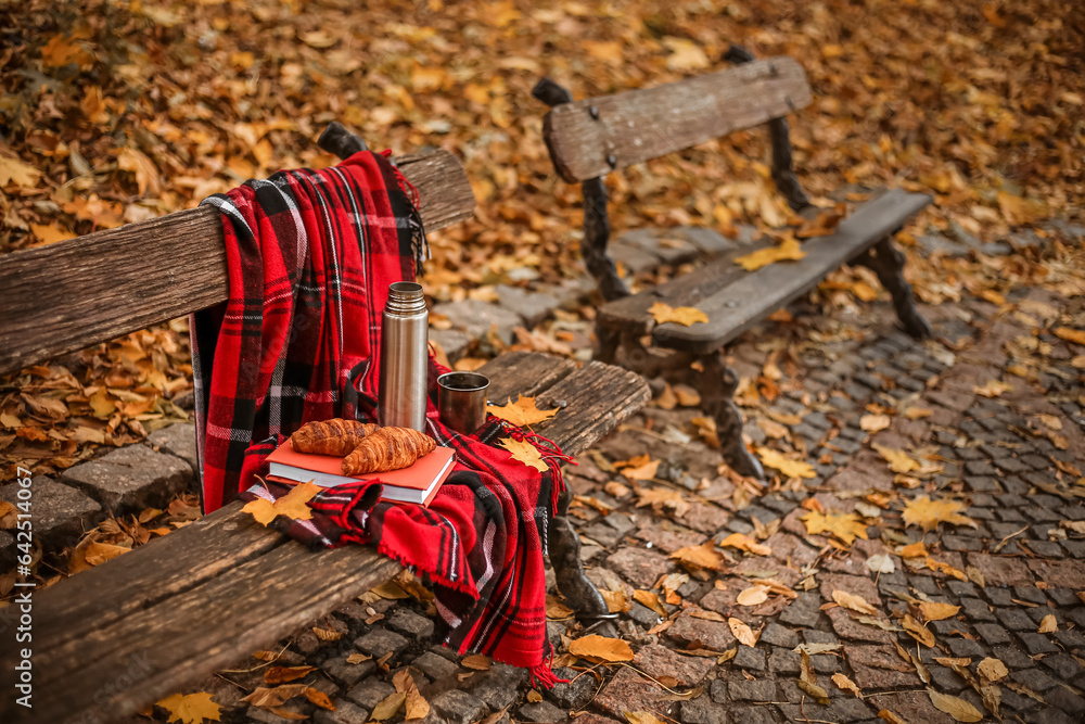 Book with croissants, thermos, cup of tea and plaid on bench in autumn park