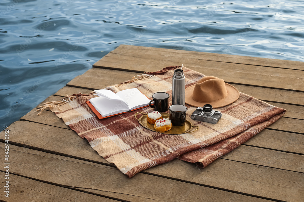 Plaid with book, cups of coffee, thermos, buns, photo camera and hat on bridge near river