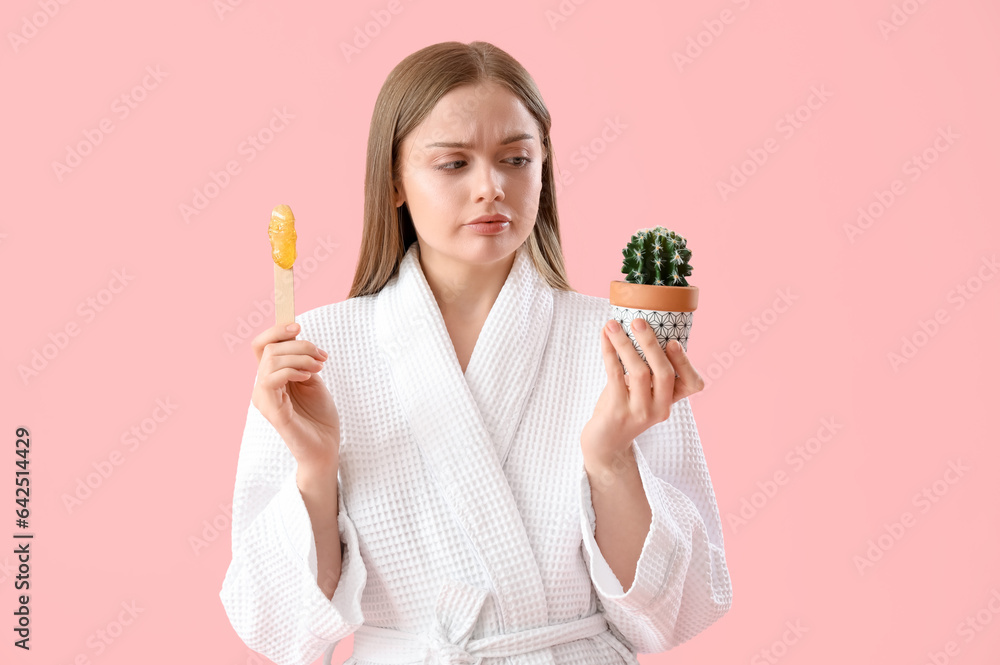 Displeased young woman holding spatula with sugaring paste and cactus on pink background