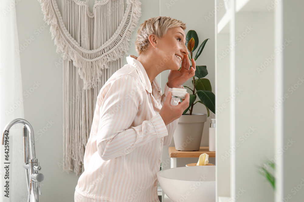 Mature woman applying facial cream near mirror in bathroom