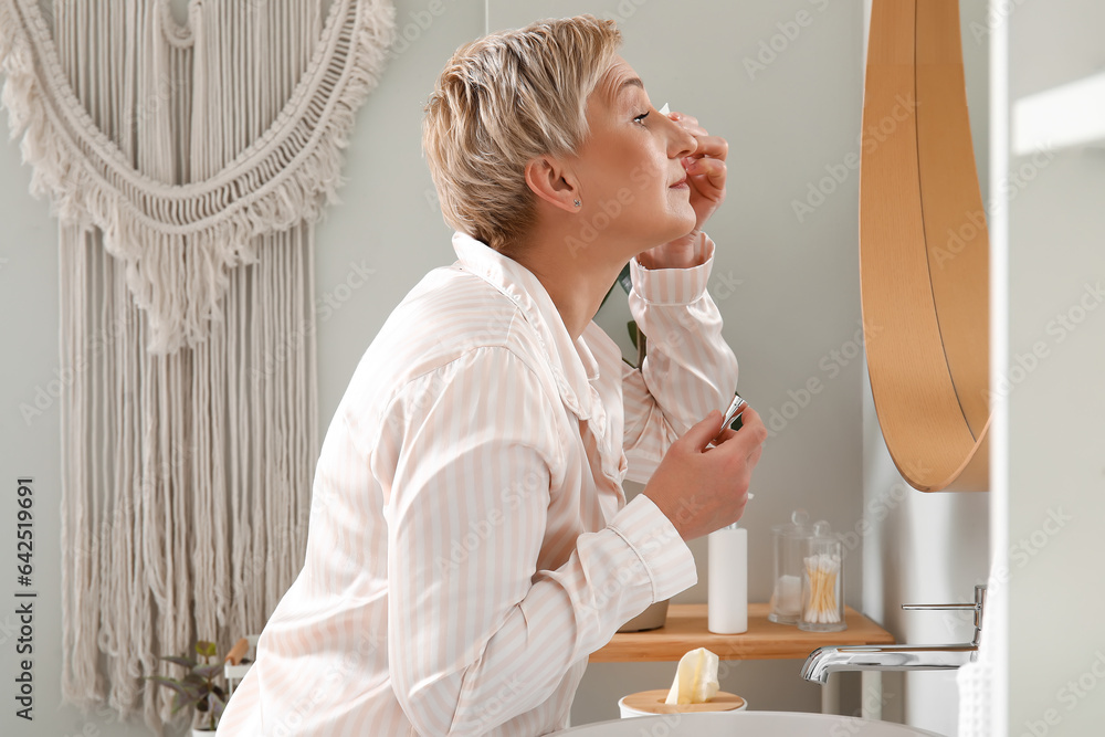 Mature woman applying under-eye cream near mirror in bathroom