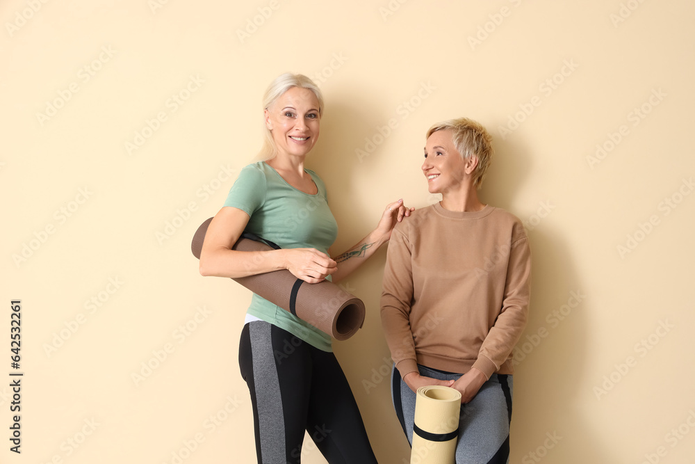 Sporty mature women with yoga mats near beige wall