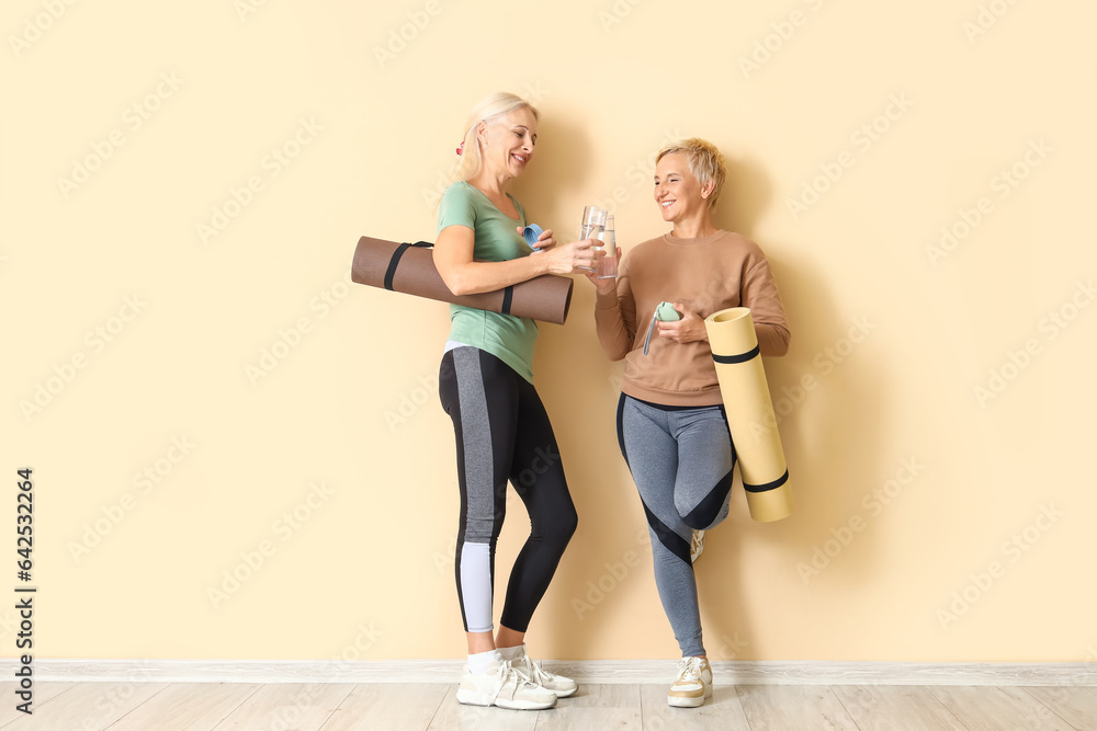 Sporty mature women with yoga mats and bottles of water near beige wall