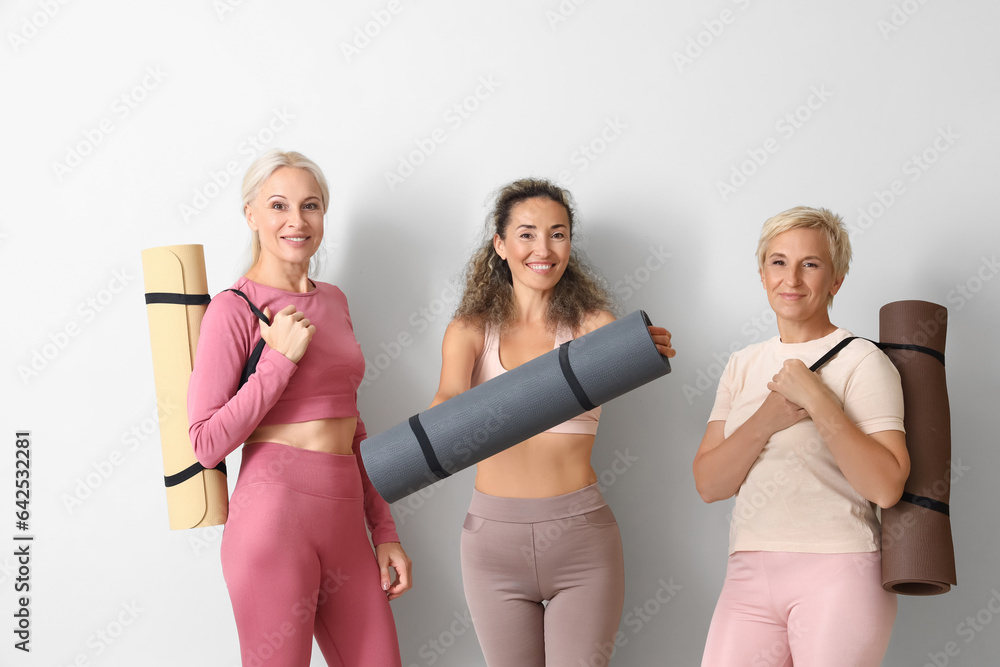 Sporty mature women with yoga mats near light wall