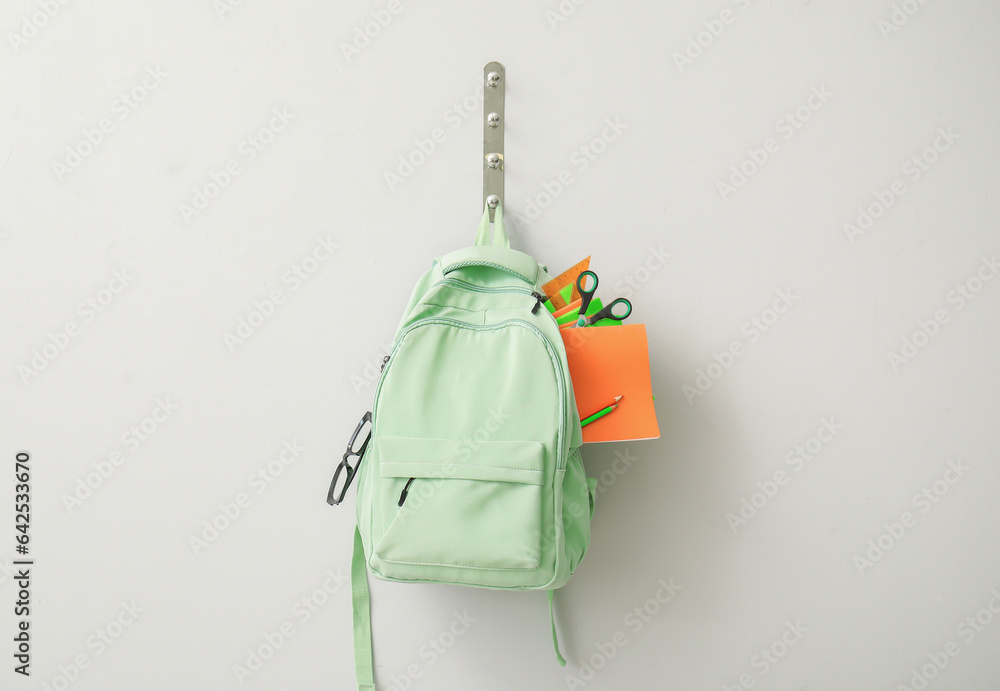 School backpack with different stationery hanging on light wall