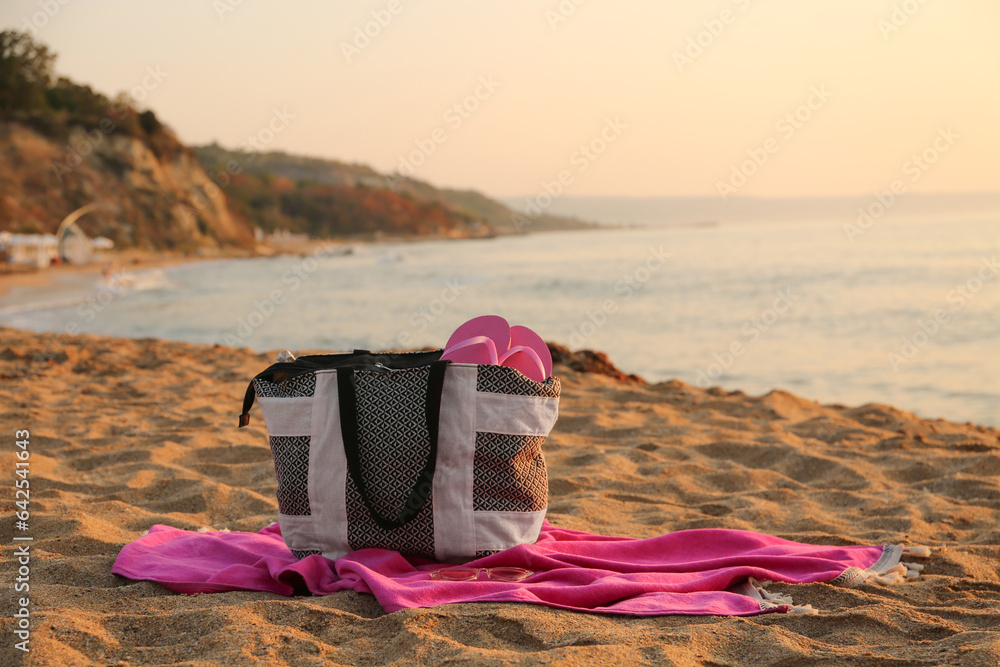 Stylish beach bag with flip-flops, sunglasses and towel on sand near sea