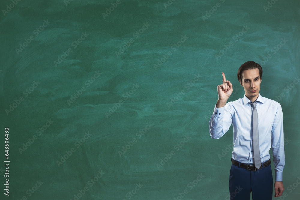 Attractive young european businessman pointing finger up while standing on empty chalkboard backgrou