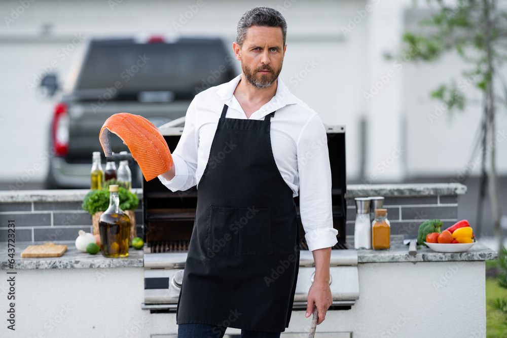 Man in chef apron hold salmon fish at barbecue grill. Male cook preparing barbecue outdoors. Bbq fis