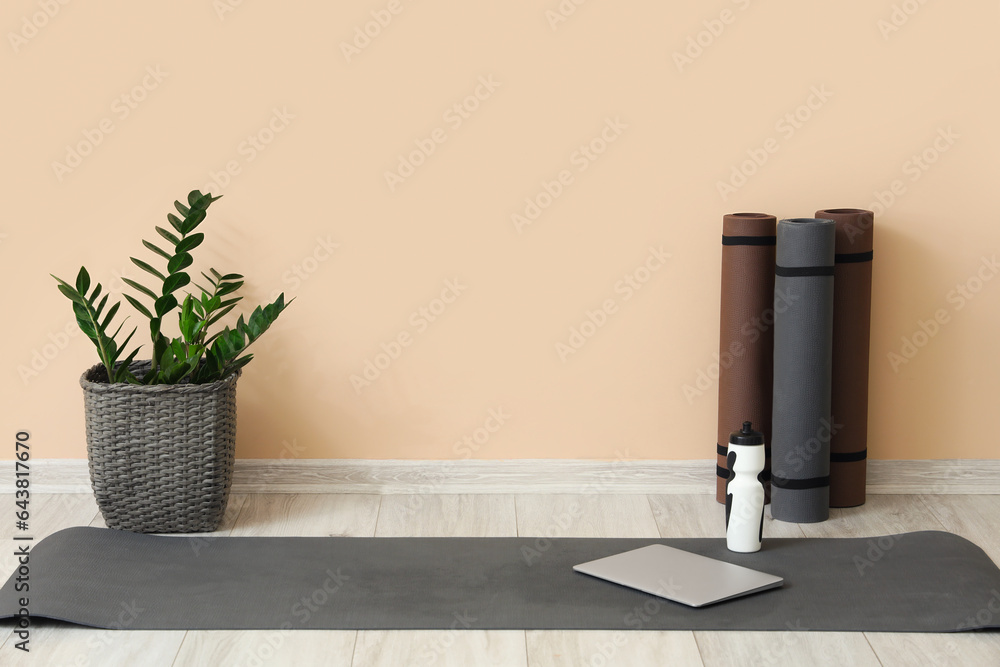 Fitness mats with bottle of water, laptop and plant near beige wall in room