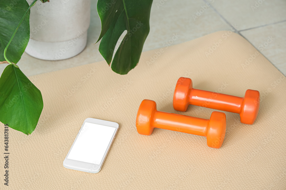 Mobile phone with dumbbells on fitness mat in gym, closeup