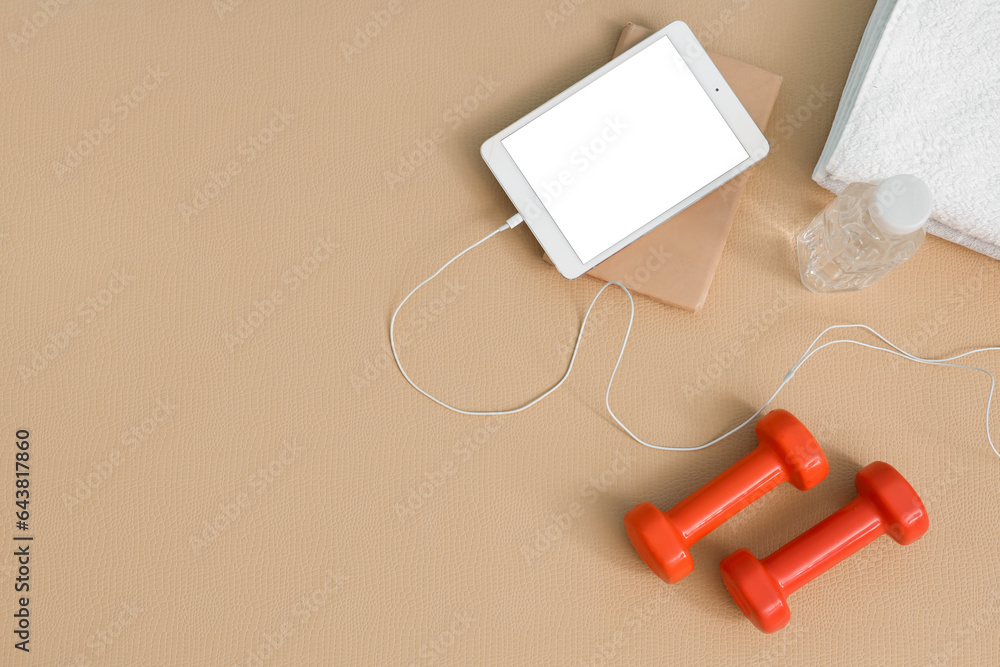 Tablet computer with dumbbells, bottle of water and towels on fitness mat in gym, top view