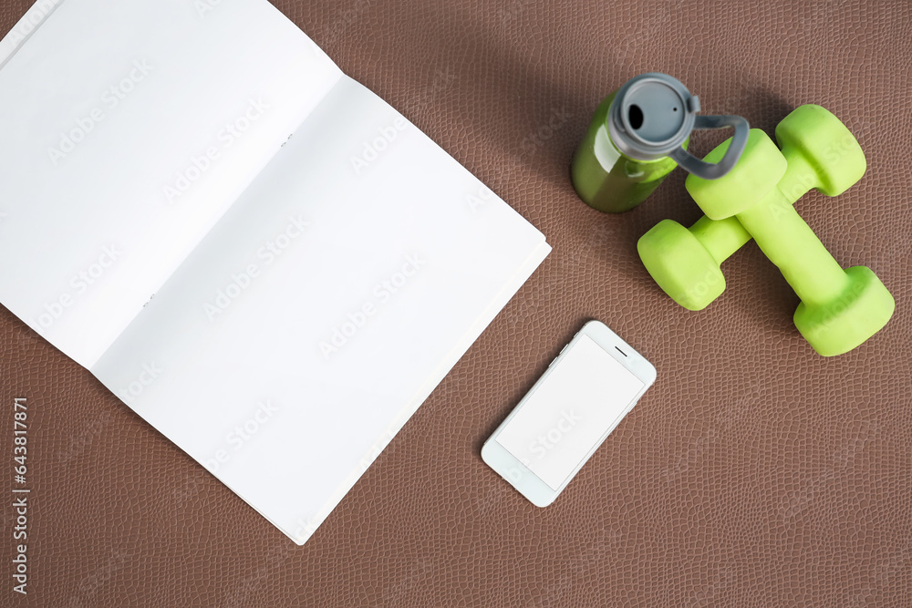 Blank magazine with mobile phone, dumbbells and bottle on fitness mat in gym, top view