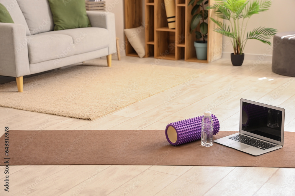 Laptop with bottle of water and foam roller on fitness mat in room