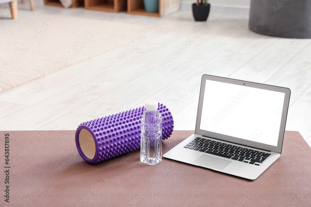 Laptop with bottle of water and foam roller on fitness mat in room