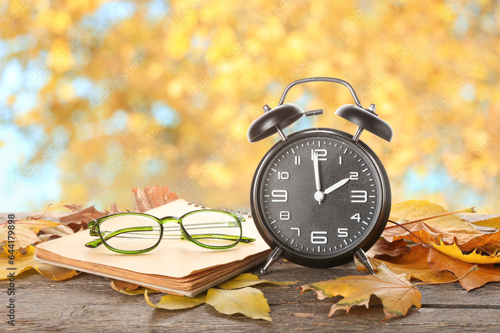 Alarm clock, notebook, glasses and autumn leaves on table outdoors