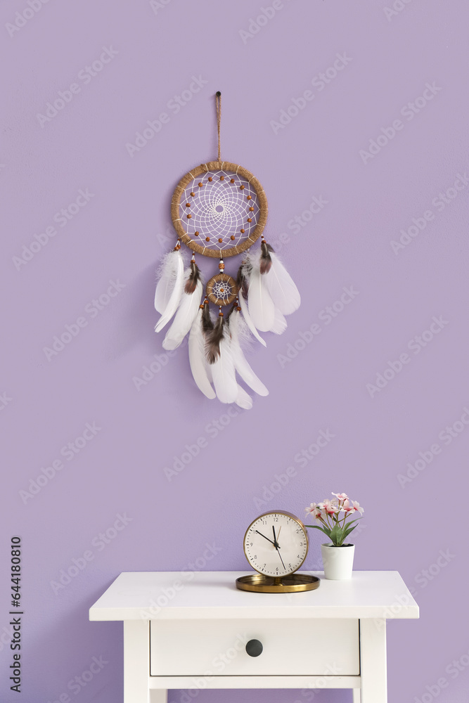 Table with alarm clock, flowers and dream catcher on lilac wall