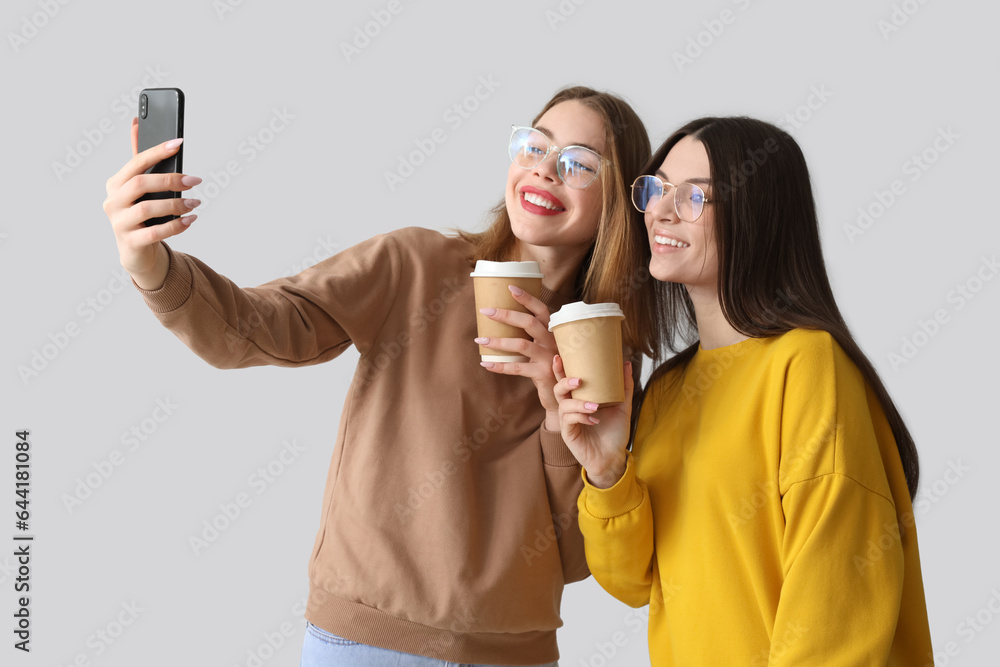 Female friends with cups of coffee taking selfie on light background