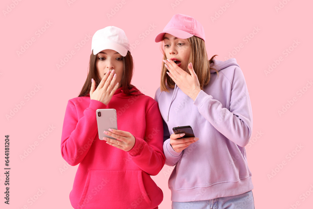 Shocked female friends using mobile phones on pink background