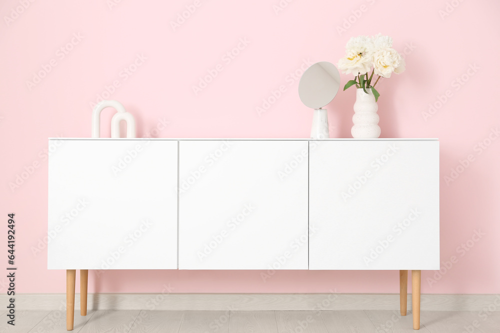 Vase of white peonies and mirror on dresser near pink wall