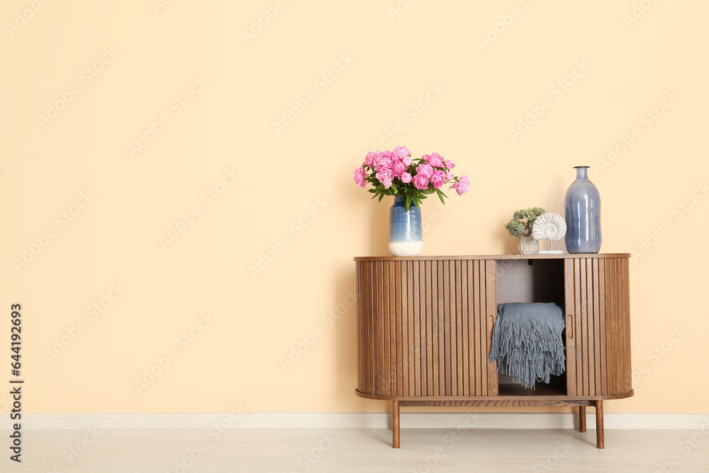 Vase of pink peonies with figurine and houseplant on dresser near beige wall