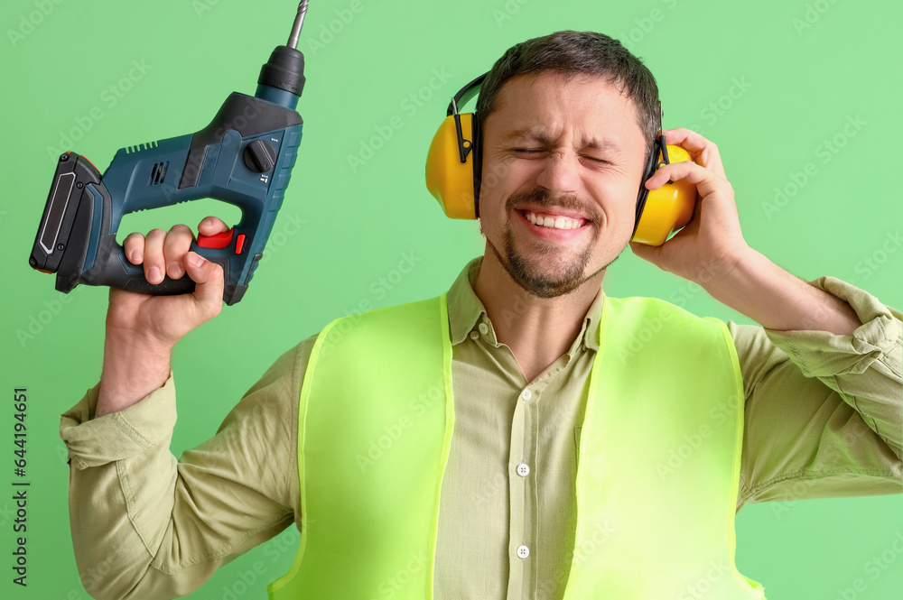 Male builder in hearing protectors with drill on green background