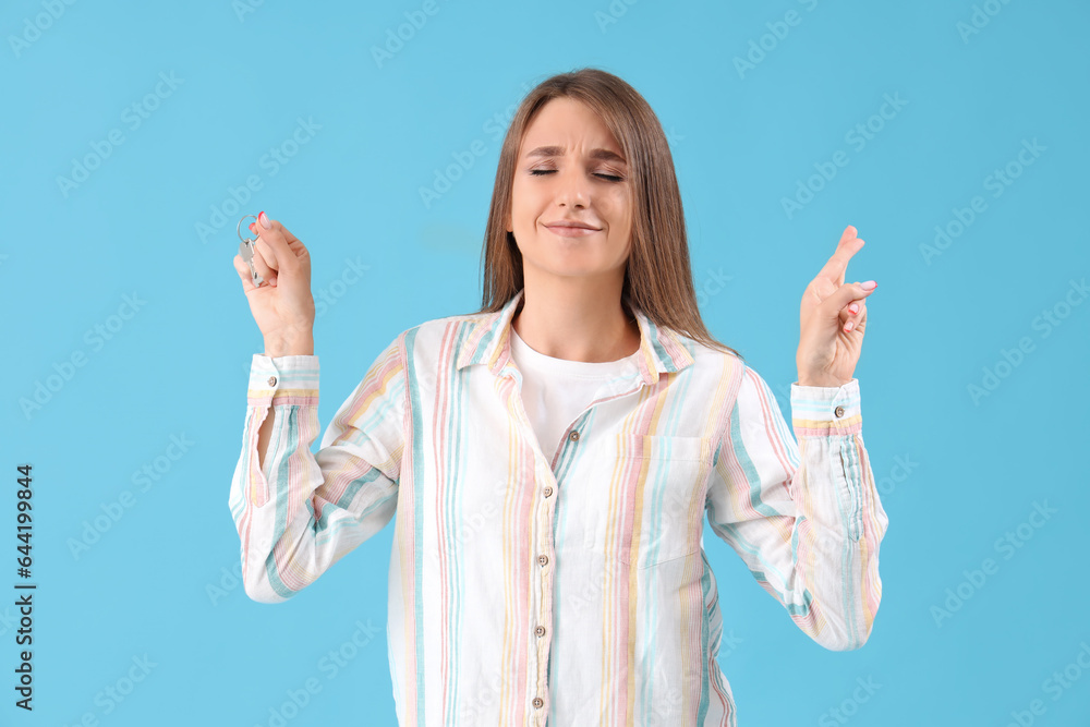 Young woman with keys from house crossing fingers on blue background