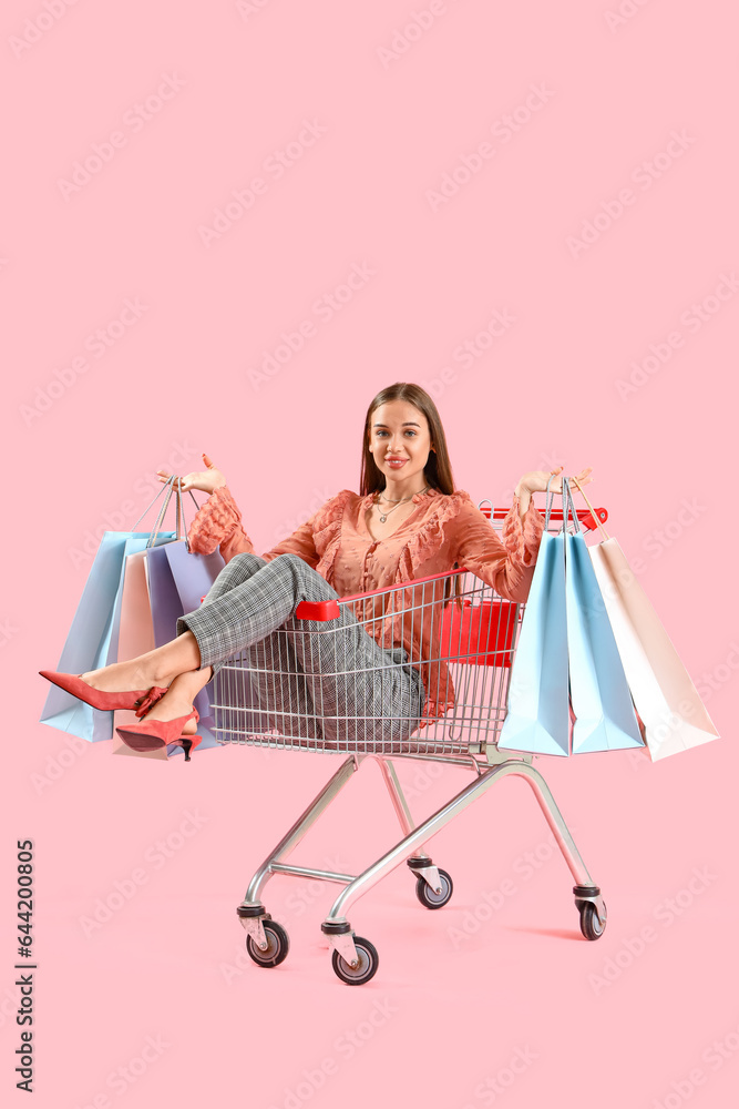 Young woman with bags in shopping cart on pink background