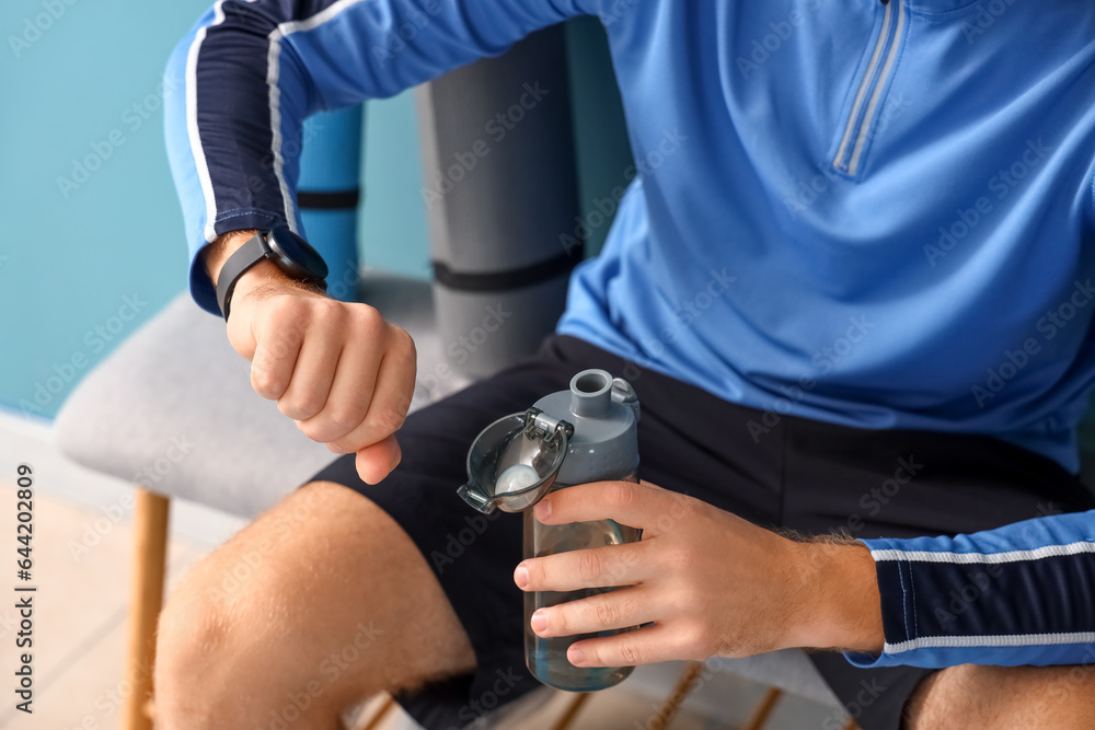 Sporty young man drinking water and looking at watch in gym, closeup